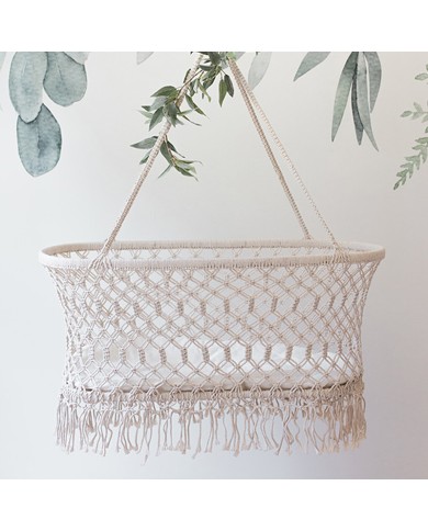 Een macramé hangwieg met aan de rand sierlijke franjes, opgehangen tegen een achtergrond van zachtgroene bladeren.