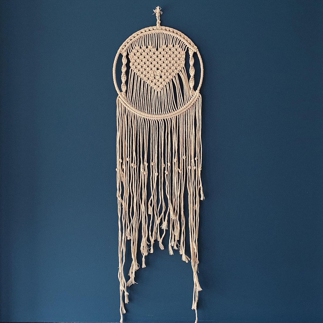 Macramé wall hanging in a heart shape with fringes, showcased against a deep blue wall.