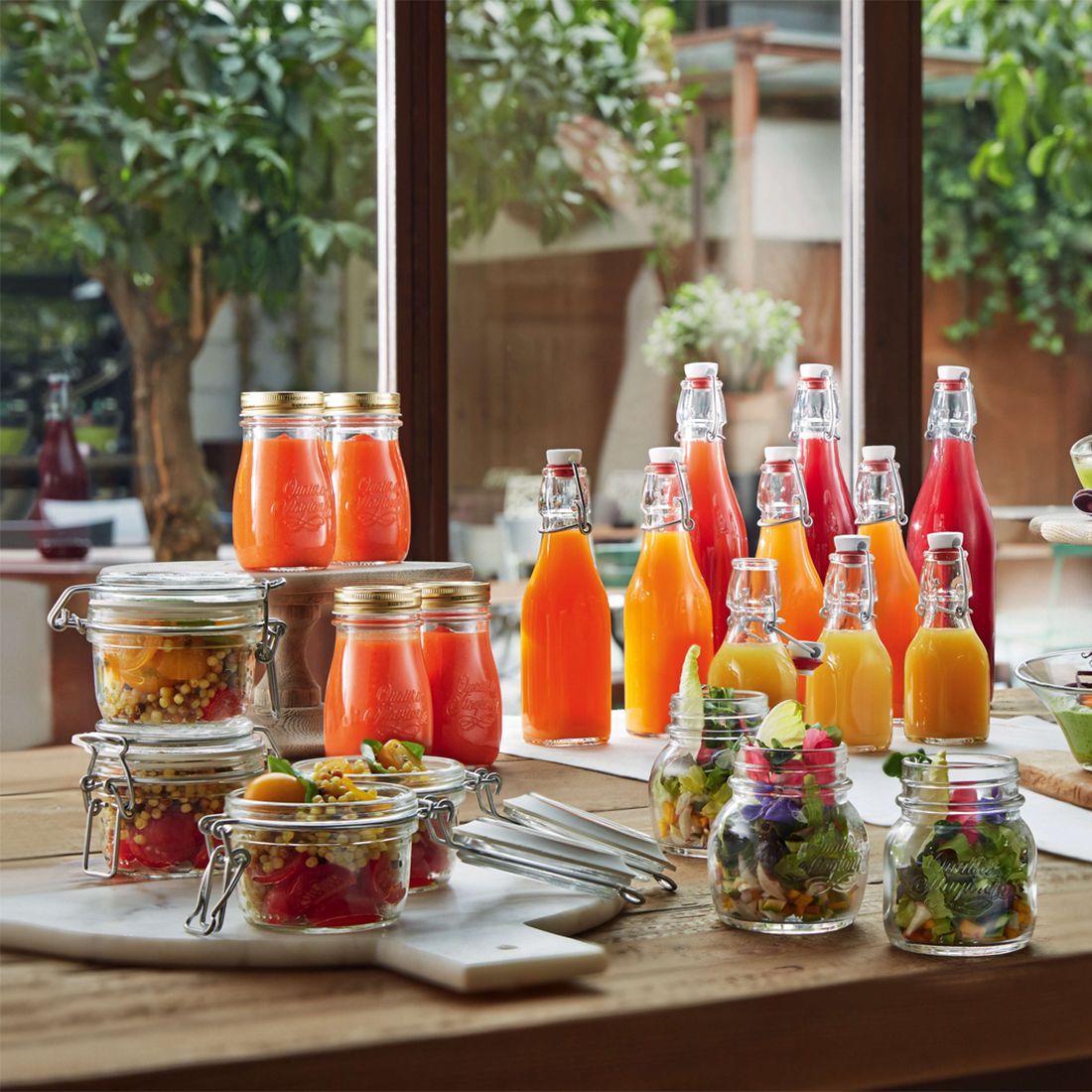 Colourful glass bottles of juice and jars of fresh salads are displayed on a wooden table.