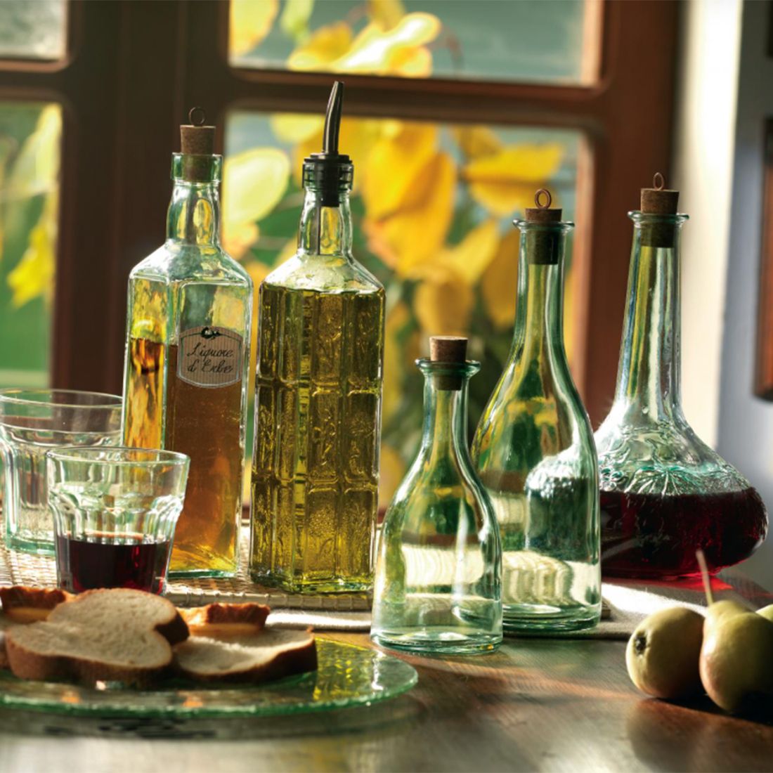 A cosy kitchen scene features green-tinted glass bottles of oil and vinegar, two glasses, bread slices, and pears.