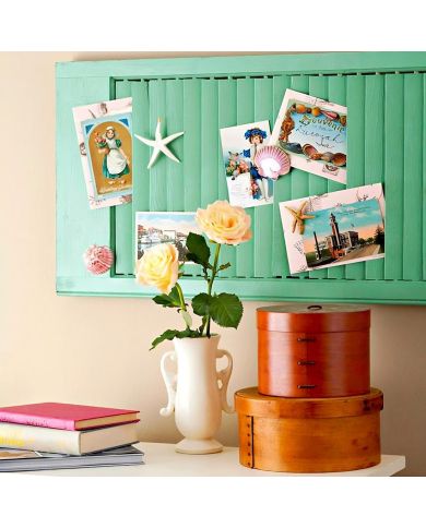 An upcycled louvre shutter displays colourful postcards and seashells, beside a white vase with yellow roses. An upcycled louvre shutter displays colourful postcards and seashells, beside a white vase with yellow roses.