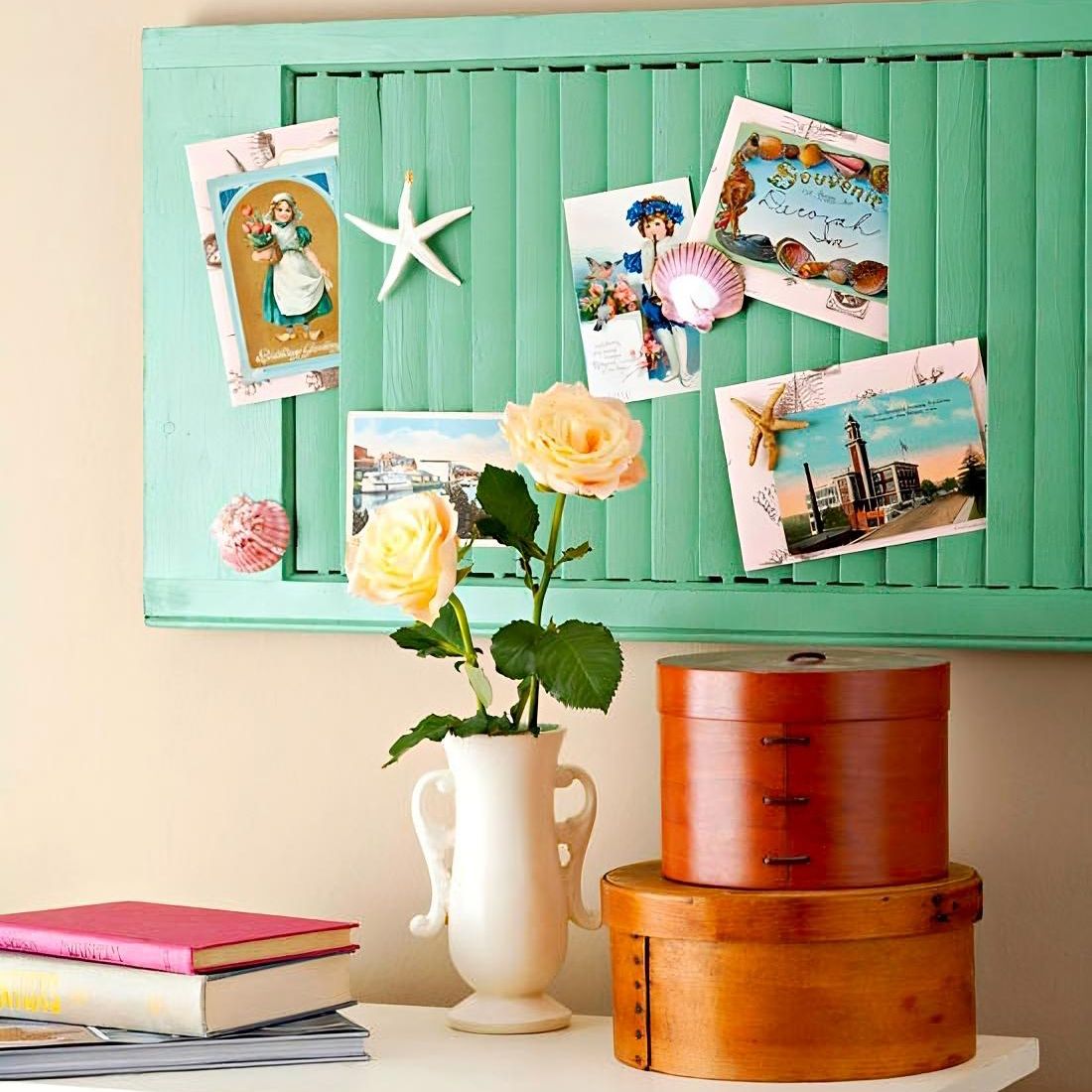 An upcycled louvre shutter displays colourful postcards and seashells, beside a white vase with yellow roses.