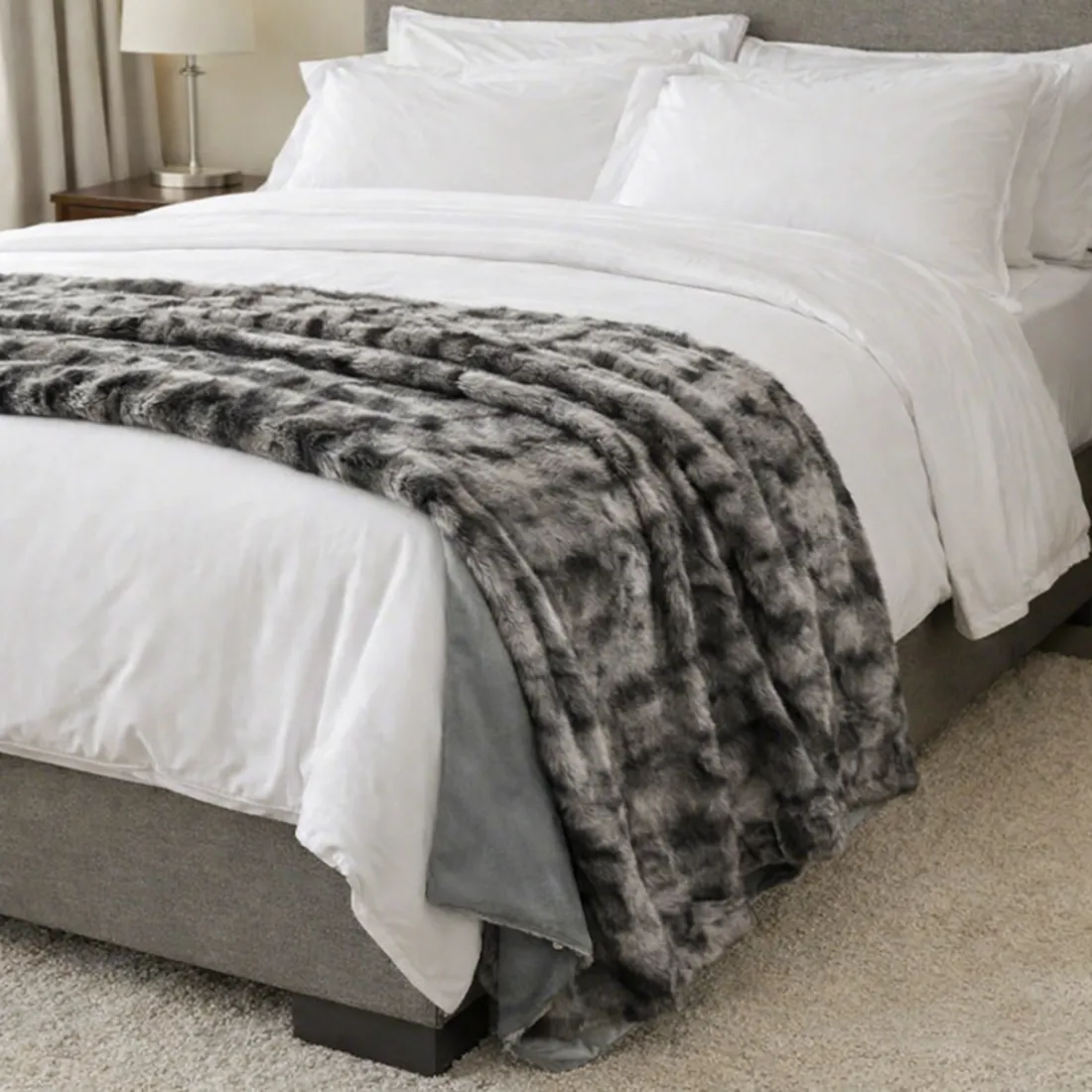 Een gezellig opgemaakt bed met witte lakens, kussens en een zilvergrijze plaid van imitatie bont in een moderne slaapkamer.
