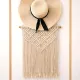A wide-brimmed straw hat with a black ribbon hangs over a macramé wall hanging featuring intricate knot designs.