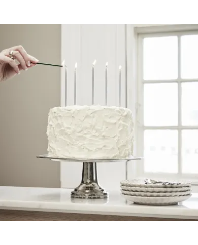 A white frosted cake placed on a silver stand, with stacked white cake plates nearby.