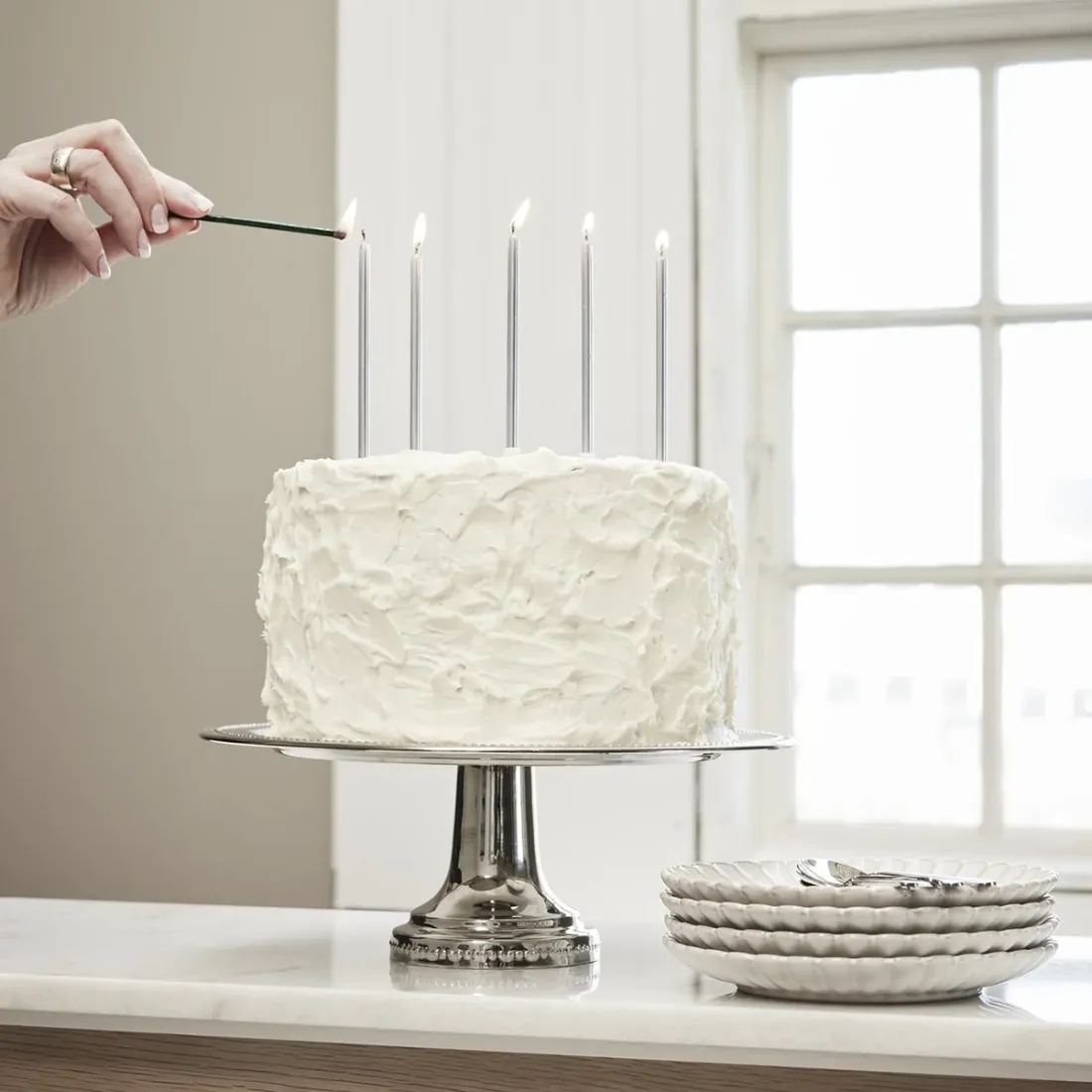 Een witte, met glazuur bedekte taart op een zilveren taartstandaard, met daarnaast opgestapelde witte gebaksbordjes.