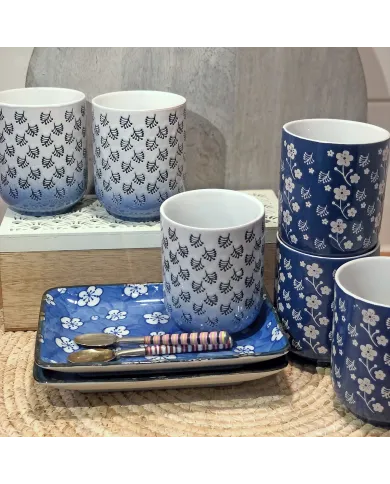 A collection of white & blue mugs and plates featuring floral designs, alongside two small spoons on a woven base.