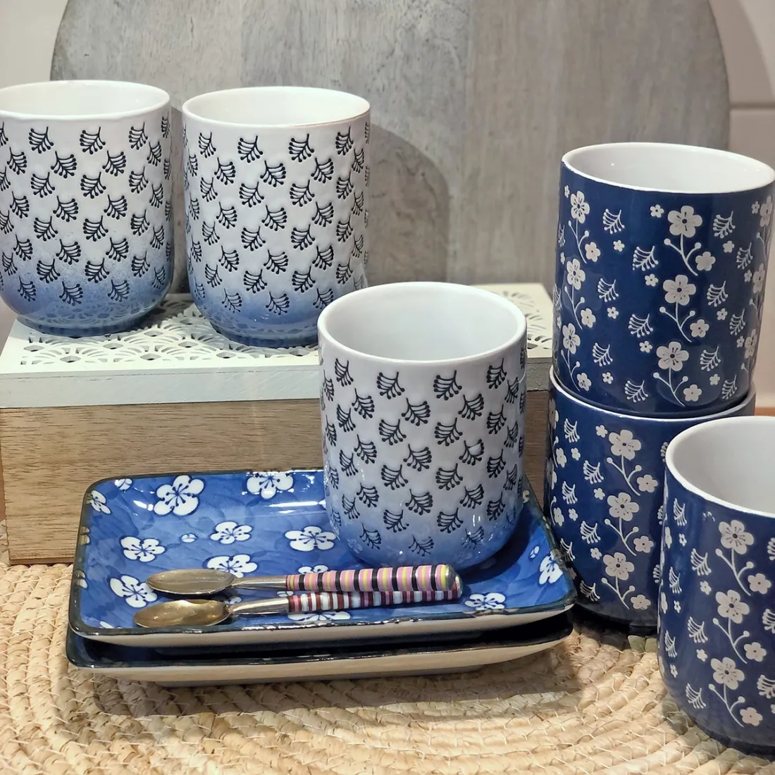 A collection of white & blue mugs and plates featuring floral designs, alongside two small spoons on a woven base.