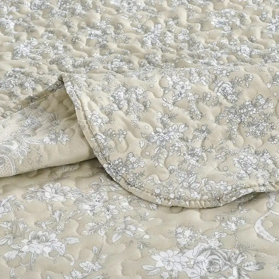 Close-up of a beige quilt, showcasing a floral toile pattern with a delicate stitching texture.