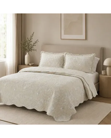 Elegant beige quilt with subtle floral print, scalloped edges and matching pillowcases on a cozy bed.