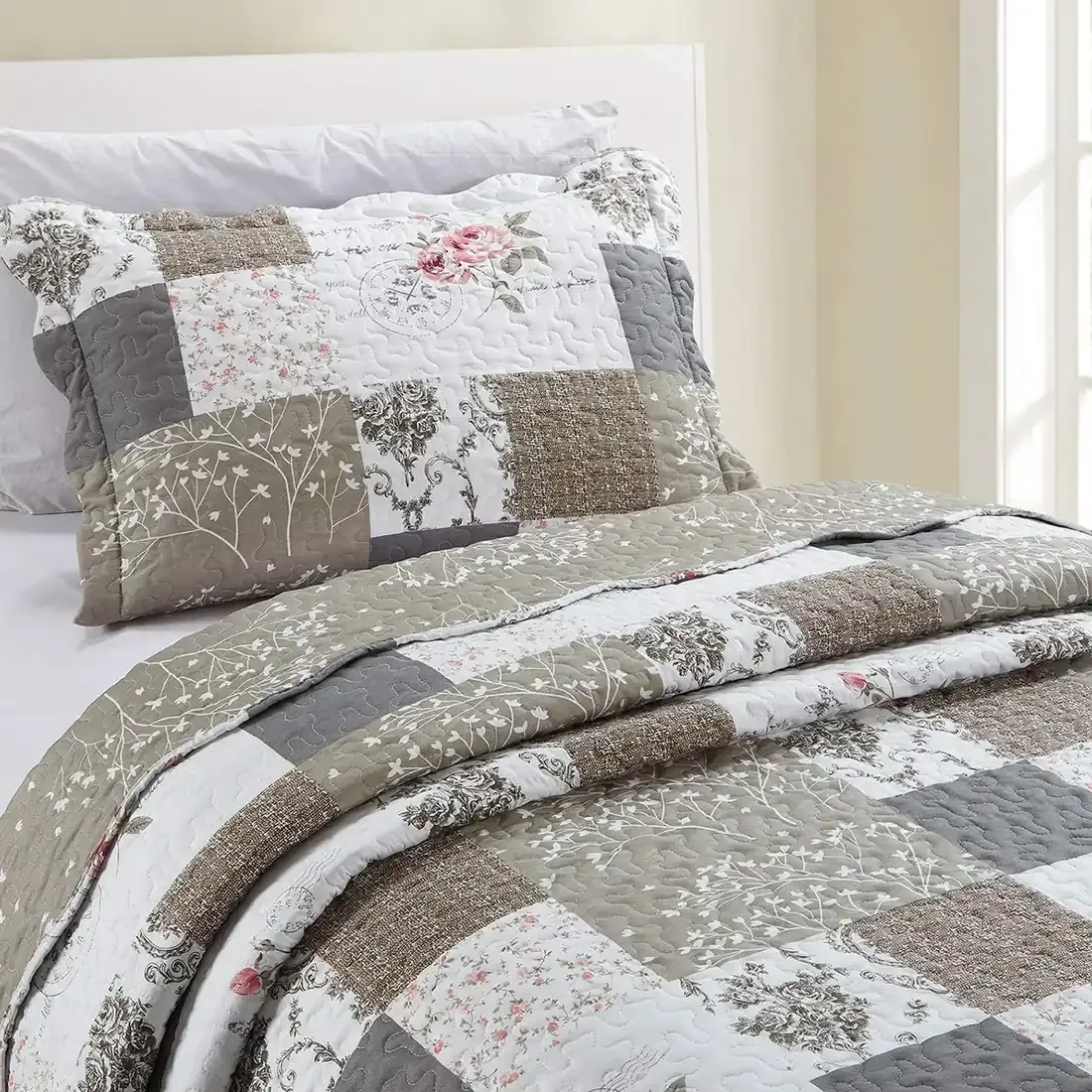 A beige patchwork pillowcase with matching bedspread on a cosy bed by a window.
