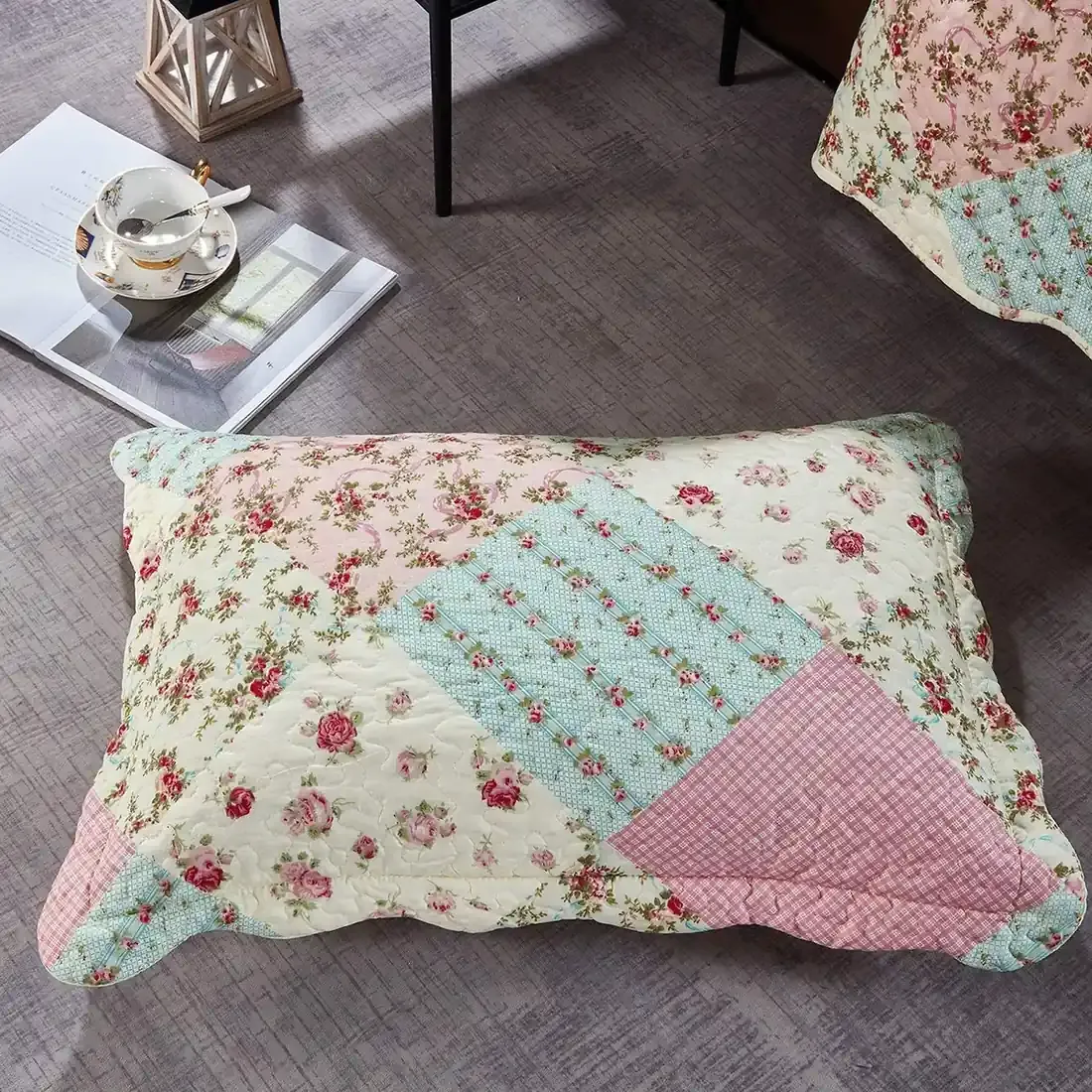 Een roze kussensloop met patchwork en lentebloemmotieven rust op een grijze vloer met structuur.