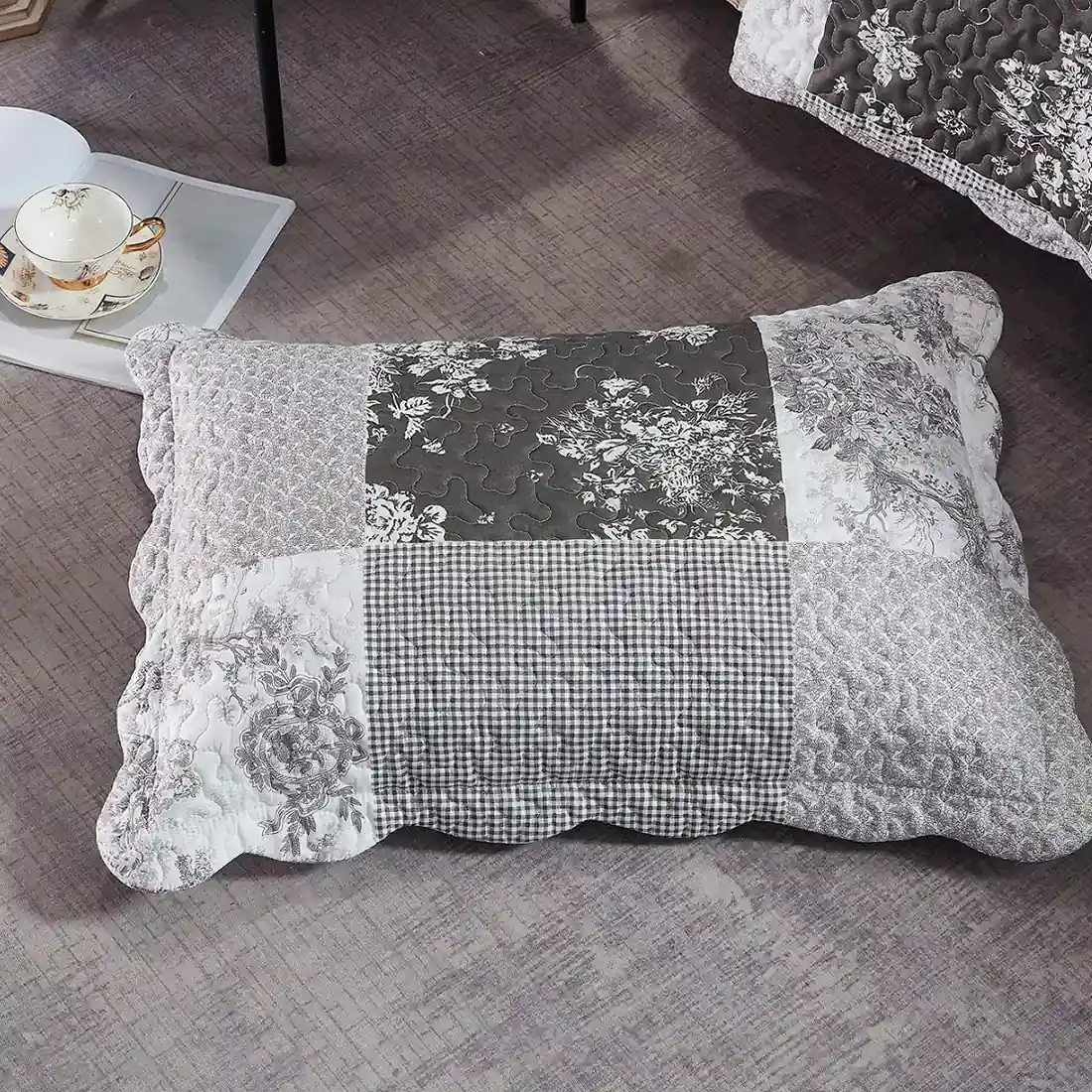 Een grijze kussensloop met Toile de Jouy motieven rust op een grijze vloer met structuur.