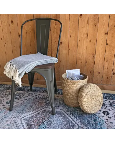 A metal chair with a grey throw blanket rests nearby a seagrass waste paper basket with lid, on a patterned rug.
