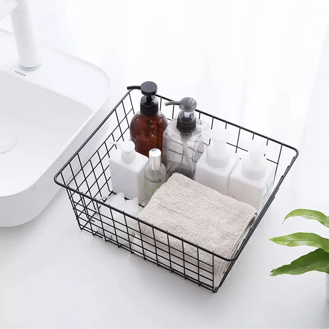 Black wire basket filled with various toiletries, a towel, and a plant in the background.