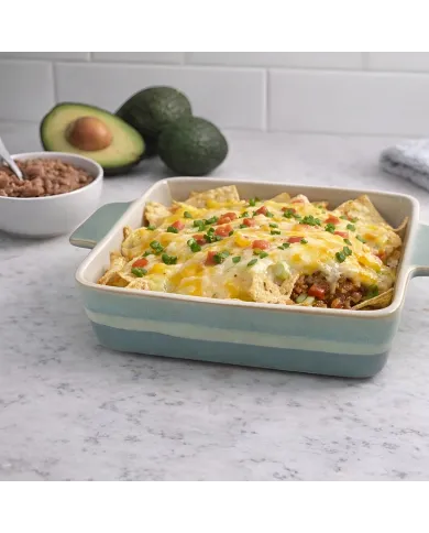 A square oven dish filled with cheesy nachos, alongside a bowl of refried beans and avocado halves.
