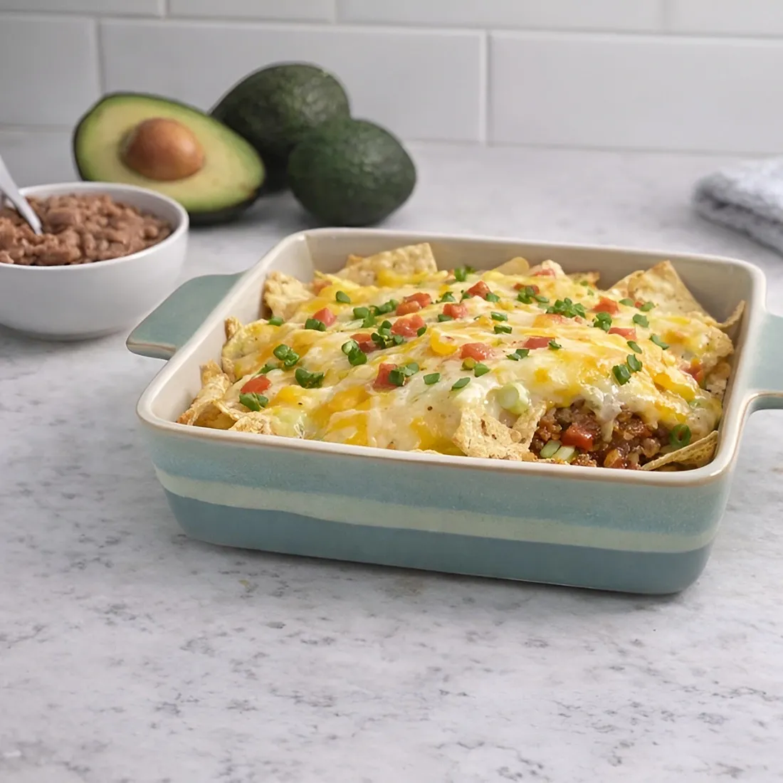 Blauwe vierkante ovenschaal gevuld met kaas overgoten nacho's, naast een kom met gehalveerde avocado's.