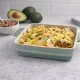 Blauwe vierkante ovenschaal gevuld met kaas overgoten nacho's, naast een kom met gehalveerde avocado's.