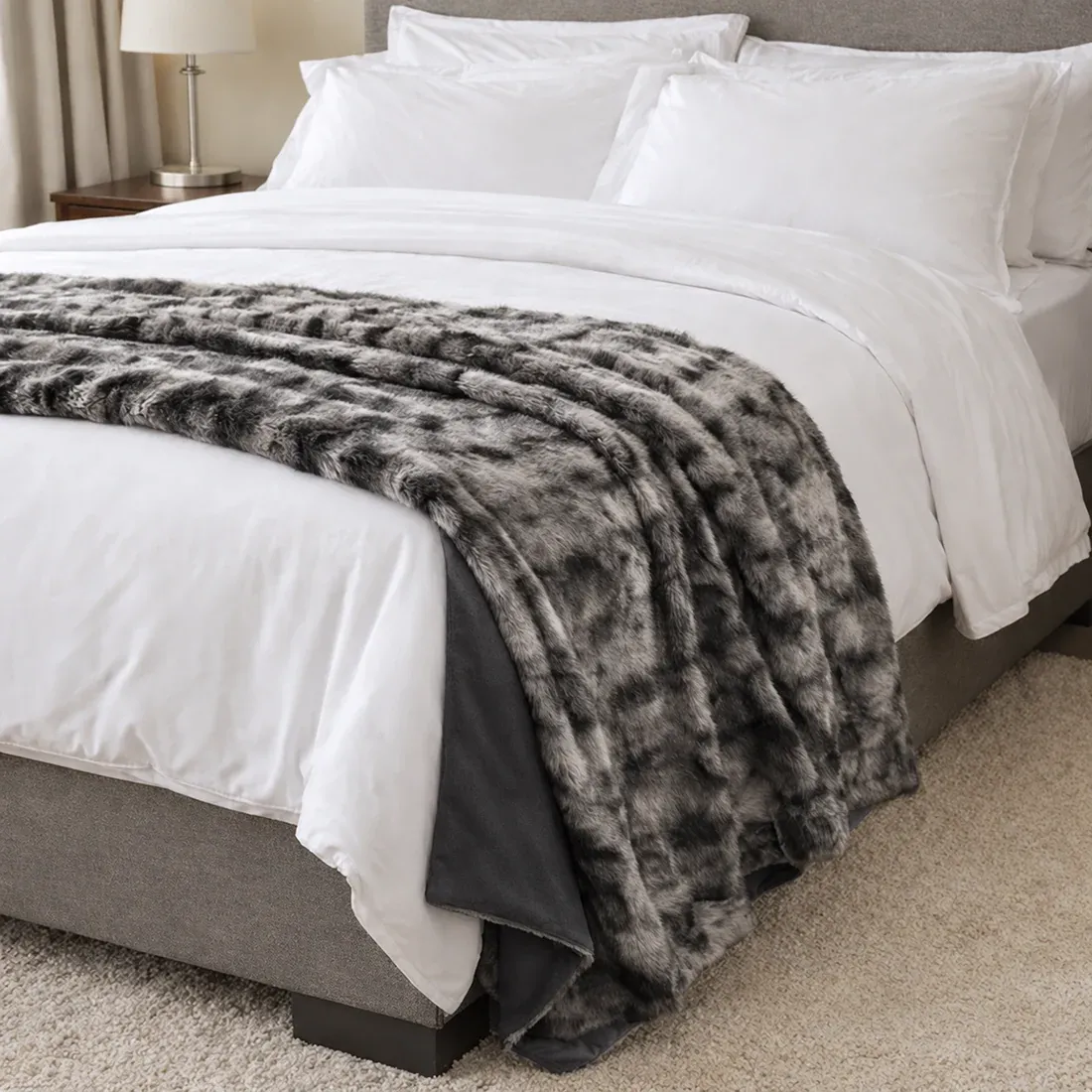 Een gezellig opgemaakt bed met witte lakens, kussens en een zilvergrijze plaid van imitatie bont in een moderne slaapkamer.