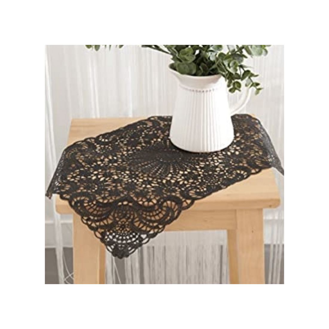 A black lace vinyl placemat on a wooden table, with a white pitcher vase filled with greenery.