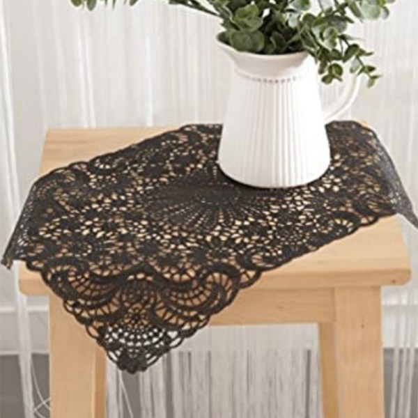 A black lace vinyl placemat on a wooden table, with a white pitcher vase filled with greenery.