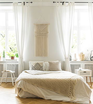 Een slaapkamer in bohemian stijl met ramen, lichte gordijnen en een macramé wandhanger boven het bed